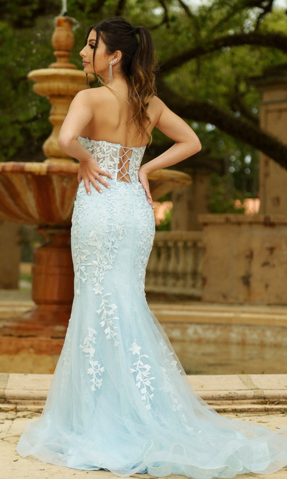 A woman stands outdoors near a stone fountain, facing away from the camera, wearing the Velvi Long Strapless Lace Mermaid Prom Dress Reva in pale blue.